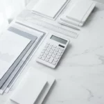 Witte rekenmachine met zilveren knoppen op marmeren bureau naast financiële documenten en grafieken in minimalistisch kantoor