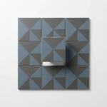 Geometrische tegels in leisteenblauw en houtskool op wit oppervlak, één tegel licht verheven met zachte schaduw en gouden accent.