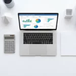 Laptop met kleurrijke inkoopdashboard grafieken op wit bureau omringd door chromen rekenmachine, notitieblok en pen in symmetrische opstelling