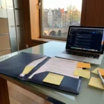 Open IT-contractmap op glazen bureau naast laptop, met sticky notes en pen, modern Amsterdams kantoor met stadszicht op de achtergrond.