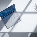 Moderne laptop met code op scherm op witte bureau, tandwiel icoon naast toetsenbord, natuurlijk licht, overhead hoek