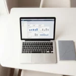 Laptop met SAP-interface op minimalistische witte bureau naast notitieboek en pen, bovenaanzicht met natuurlijk licht
