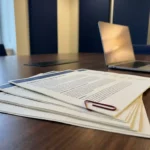 Stapel softwarelicentiedocumenten op een bureau, één contract gemarkeerd met een rode paperclip, laptop op de achtergrond.