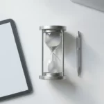 Zilveren zandloper met stromend zand op wit bureau naast digitale tablet en geometrische pen, minimalistisch design