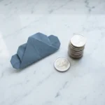 Origami wolkfiguur naast een stapel euromunten op wit marmer, één munt apart gelegd, minimalistische bovenaanzichtfoto.