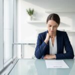 Professionele zakenvrouw in marineblauw jasje bekijkt cv-document aan moderne glazen bureau in wit kantoor